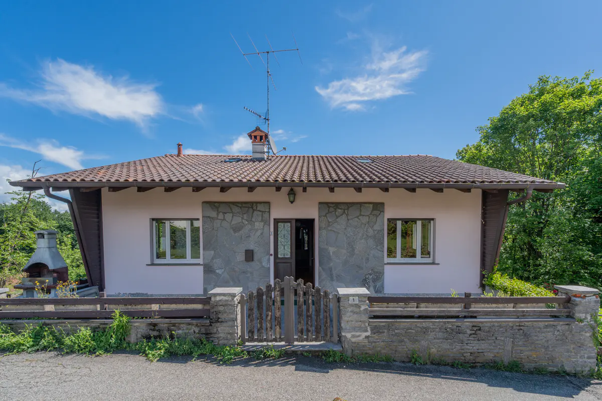 Single-story house with light pink walls, stone accents, a tiled roof, and a wooden gate under a bright blue sky.