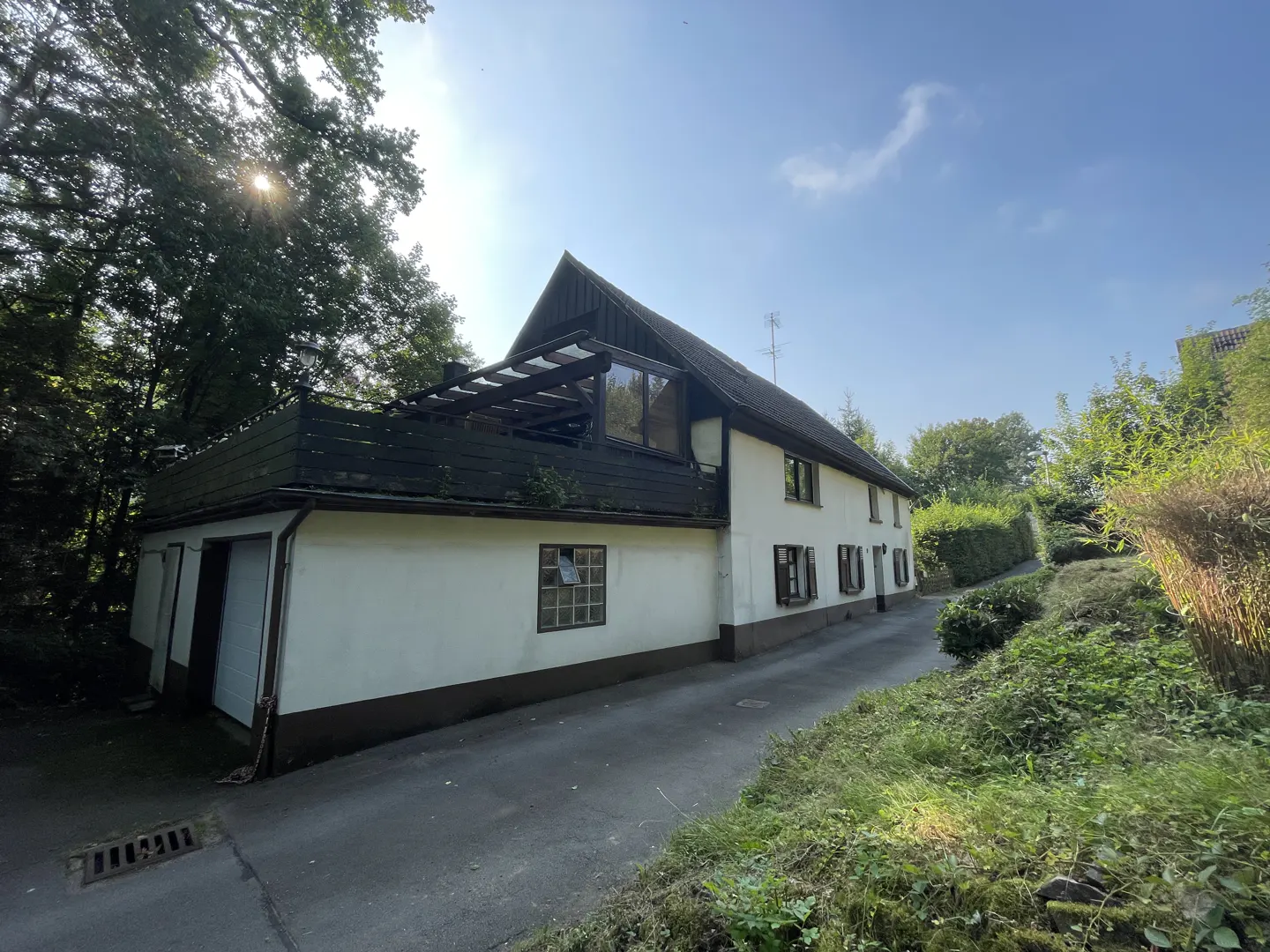 Exterior view of a two-story white house with dark trim, a balcony, and a driveway surrounded by greenery.