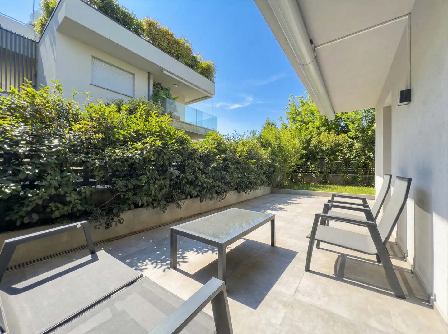 Outdoor patio with gray tile, table, and chairs. Green bushes line the back, with a white building and blue sky in the background.