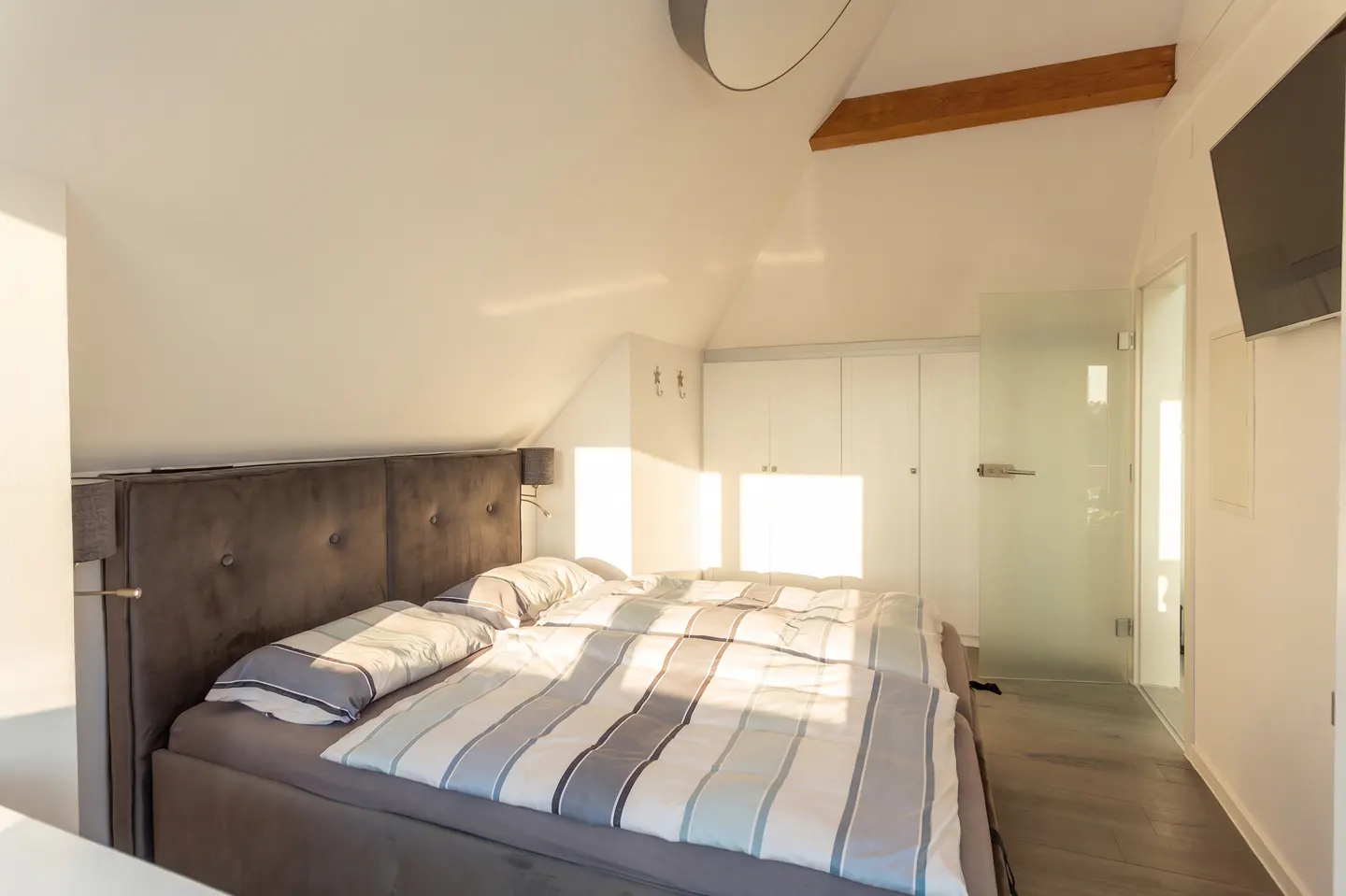 Bright bedroom with a gray upholstered bed, striped bedding, and white walls. A glass door leads to an en-suite bathroom.