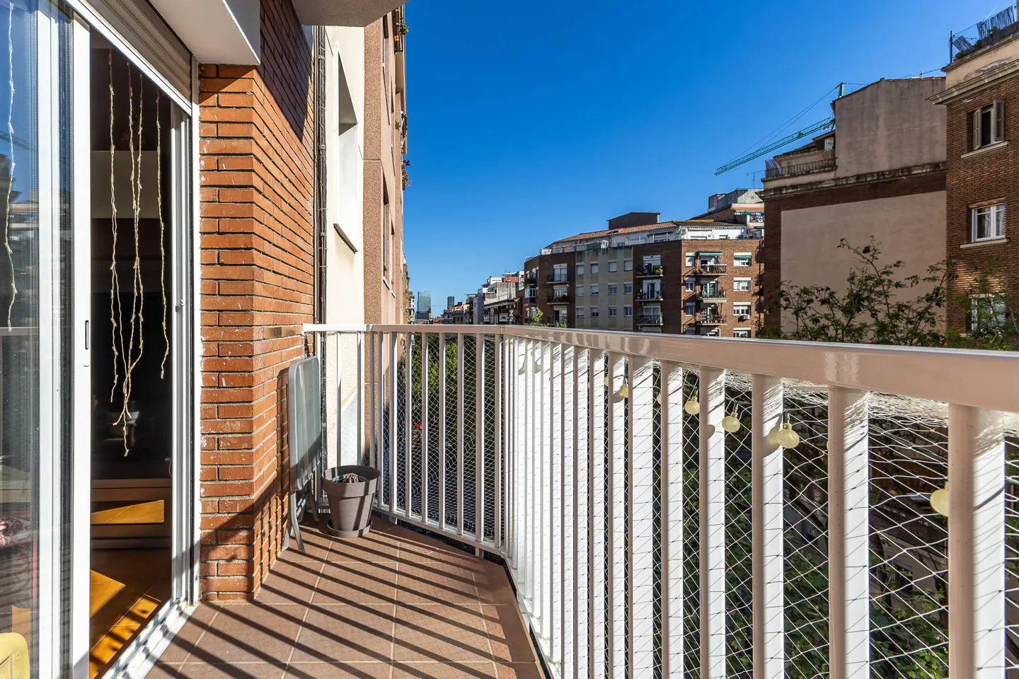 Balcony with white railings, brown tile floor, and brick wall. A pot sits near a folding table. City buildings are visible in the background.