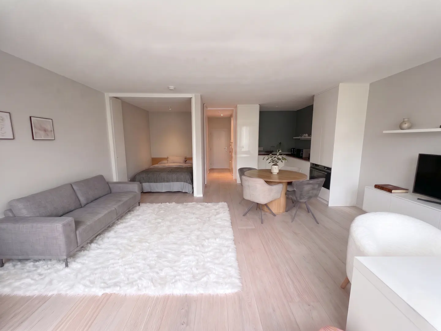 A modern apartment interior with a gray sofa, white rug, round table with chairs, and a bedroom alcove.