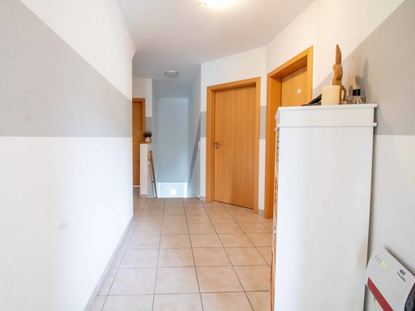 Hallway with beige tile floor, white and gray striped walls, and wooden doors. A white cabinet with decor sits on the right.