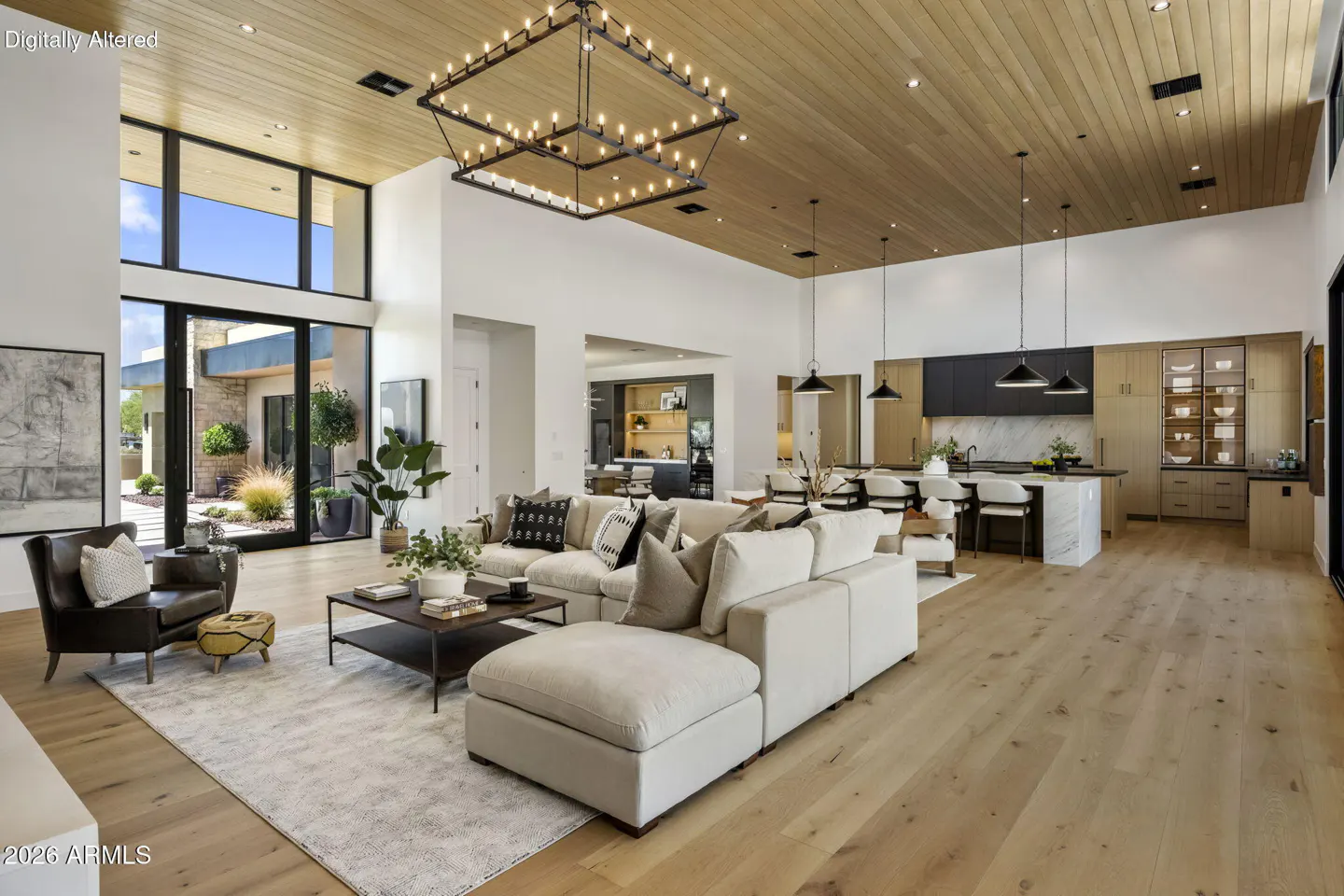 Open-concept living room with a beige sectional, wood floors, and a modern chandelier. Kitchen island and cabinets are visible in the background.