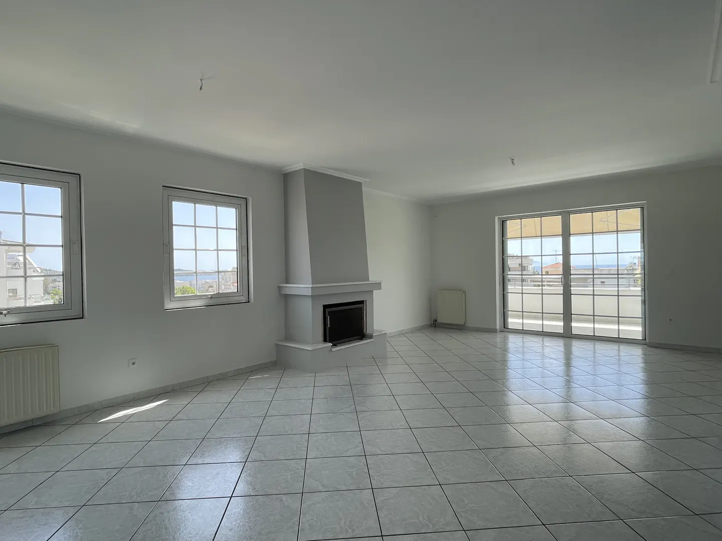 Bright, empty room with white walls, tile floor, and a gray fireplace. Windows and sliding doors offer a view of the outside.