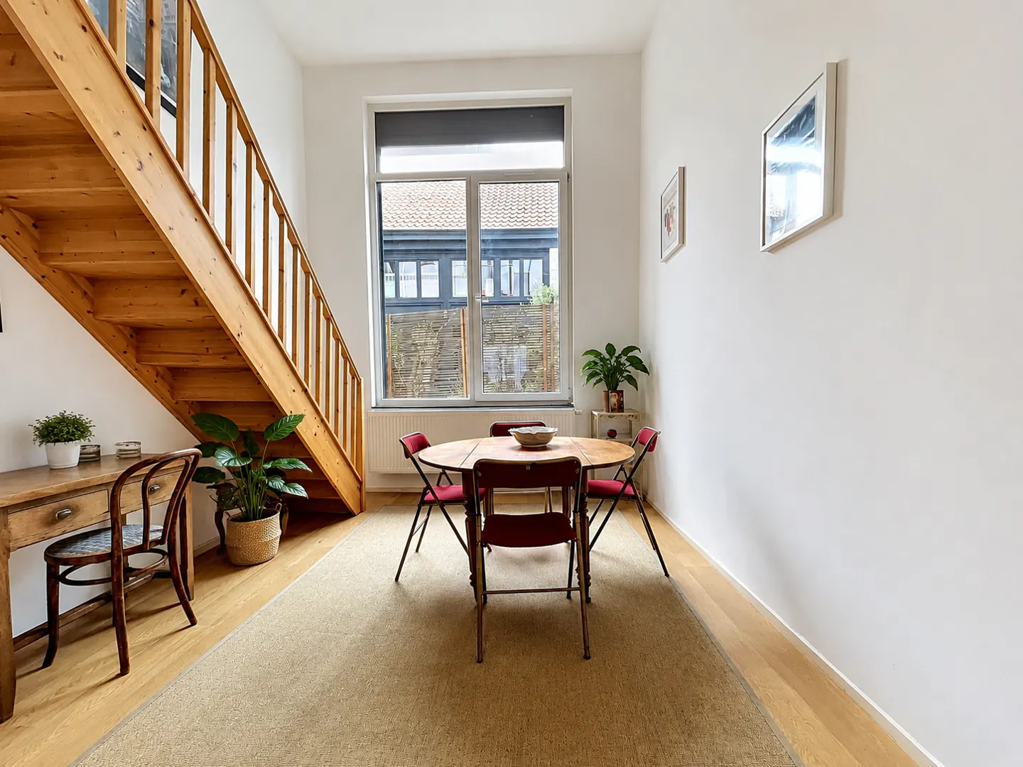 Bright room with wooden stairs, table with red chairs, and a desk. A large window shows a building outside. Walls are white.