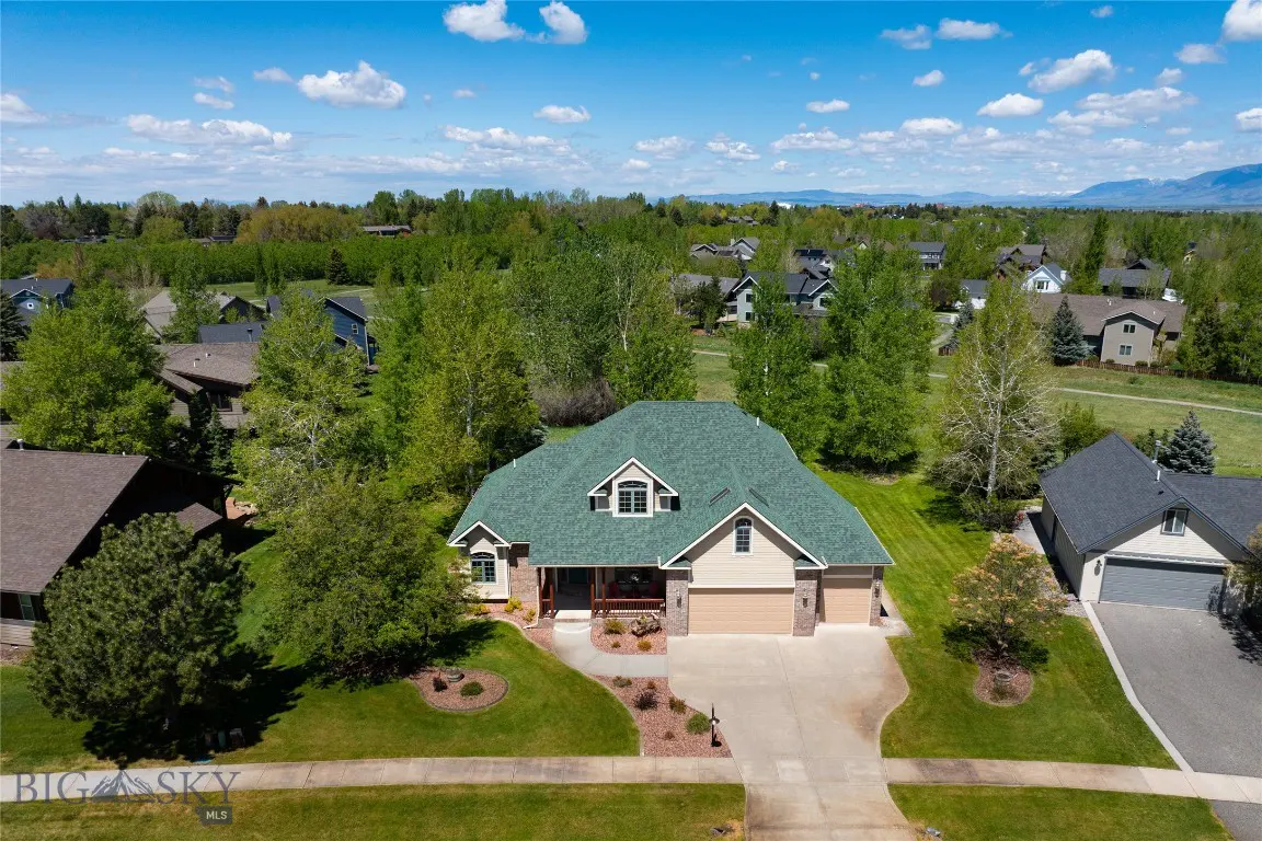 Aerial view of a beige house with a green roof, surrounded by trees and a blue sky with clouds.