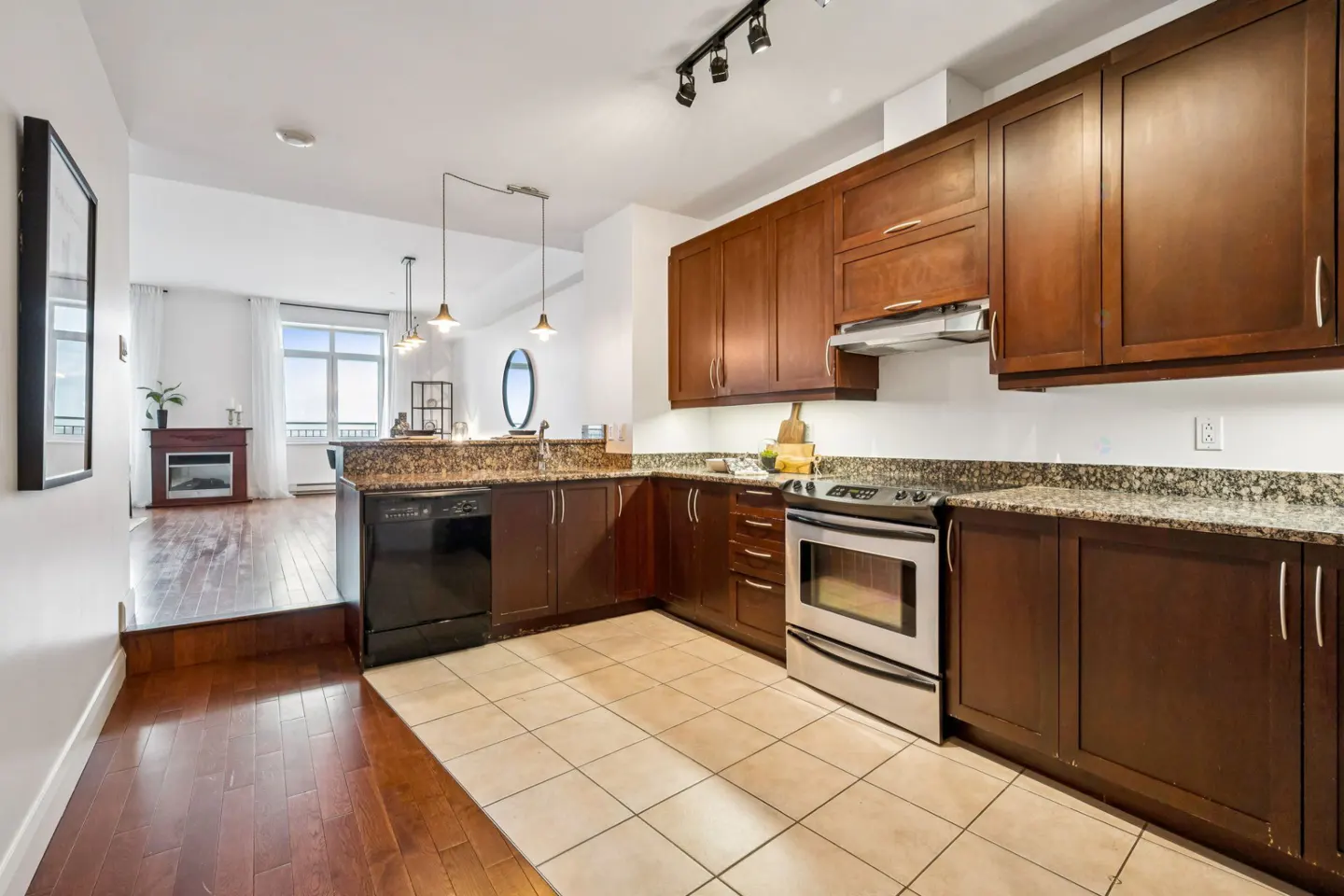 A kitchen with brown cabinets, granite countertops, and stainless steel appliances. The floor is tiled, and there is a living room in the background.