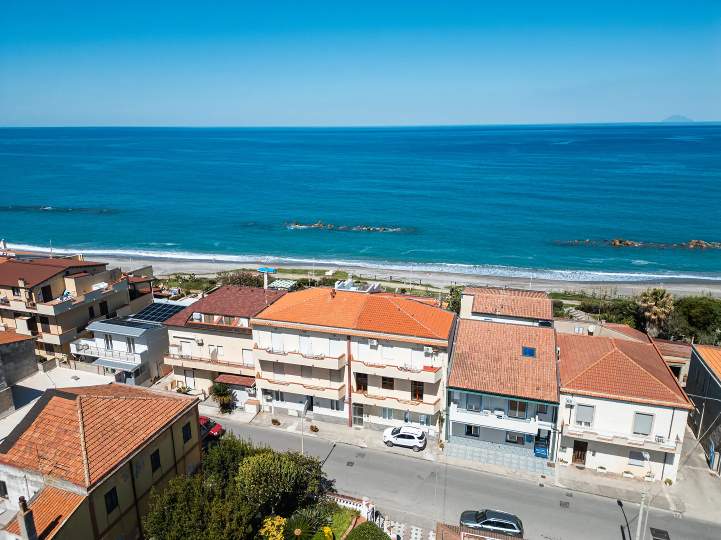Coastal view of houses with orange tile roofs, a sandy beach, and a blue ocean under a clear sky.