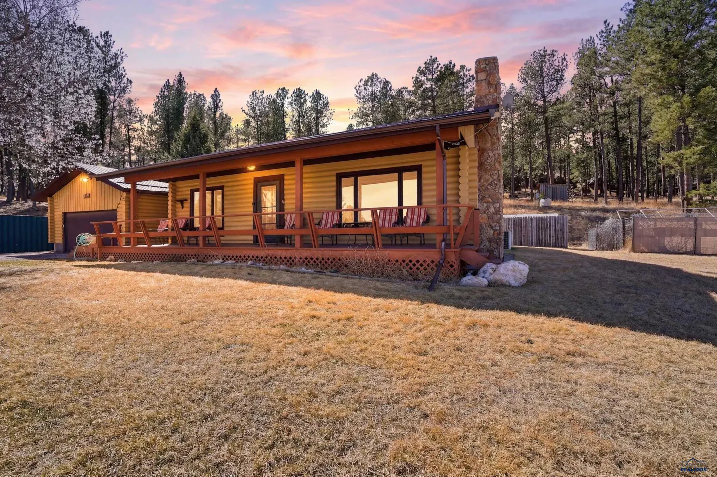 Exterior view of a log cabin home with a stone chimney and a wooden porch with chairs. The sky is pink and orange.