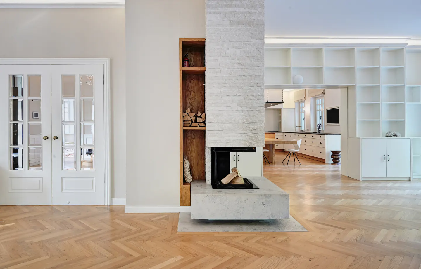 Bright, modern living room with herringbone wood floors. A white brick fireplace divides the space, with a wood shelf on one side and a kitchen visible beyond. White double doors on the left.
