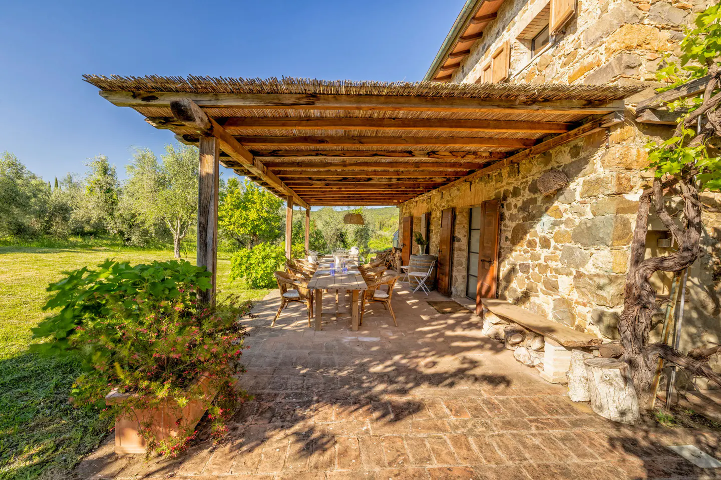 Outdoor patio with a long wooden table and chairs under a thatched roof, attached to a stone building.