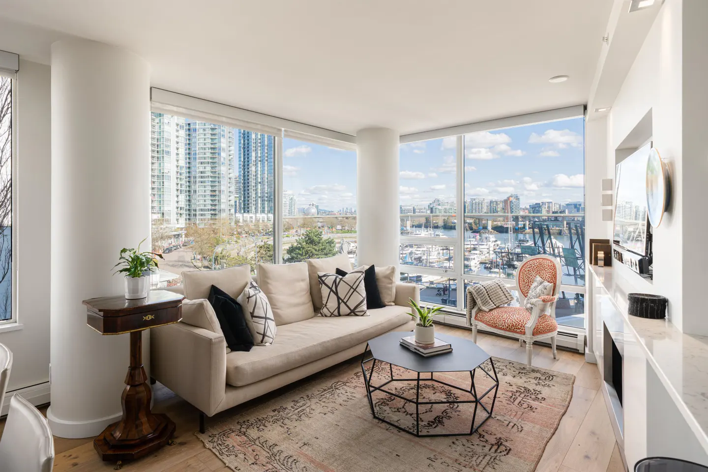 Bright living room with beige sofa, patterned pillows, and geometric coffee table on a vintage rug. Large windows offer city and harbor views.
