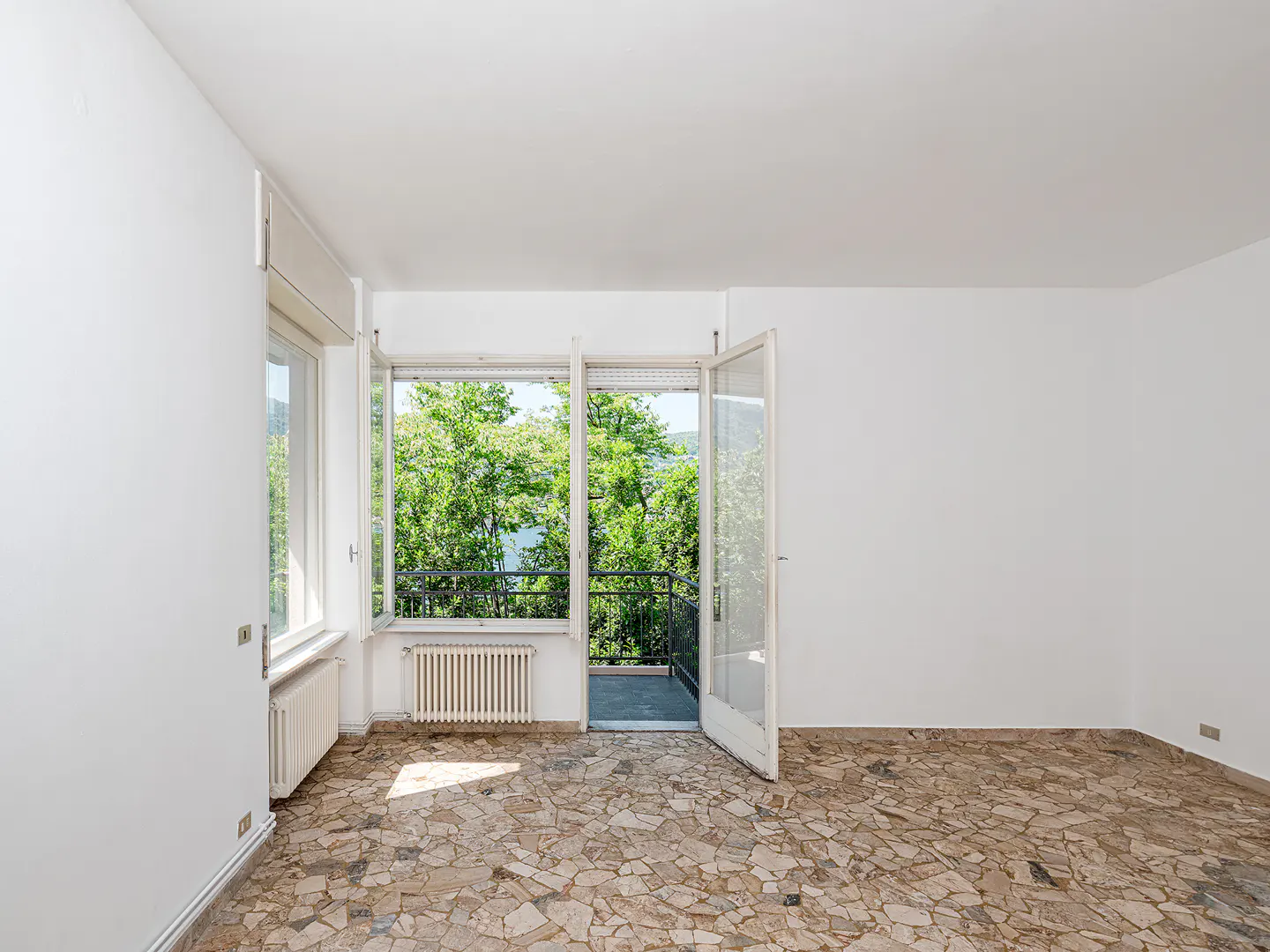 Empty room with white walls, stone tile flooring, and an open door leading to a balcony with lush green trees.