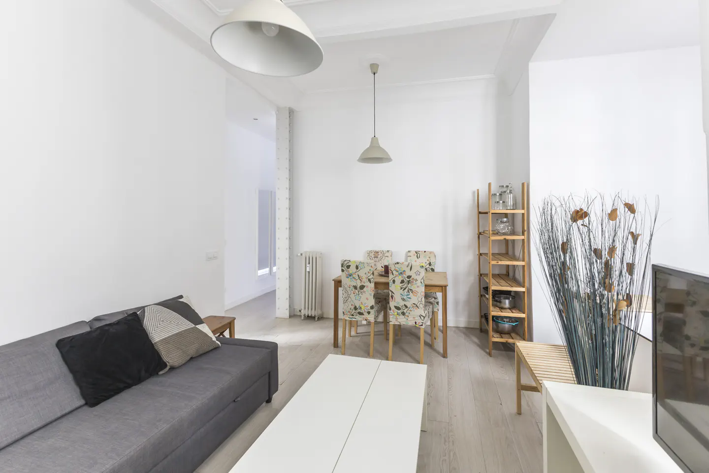 Bright living room with a gray sofa, white coffee table, and wooden dining set with floral chairs. A tall plant and wooden shelf add decor.