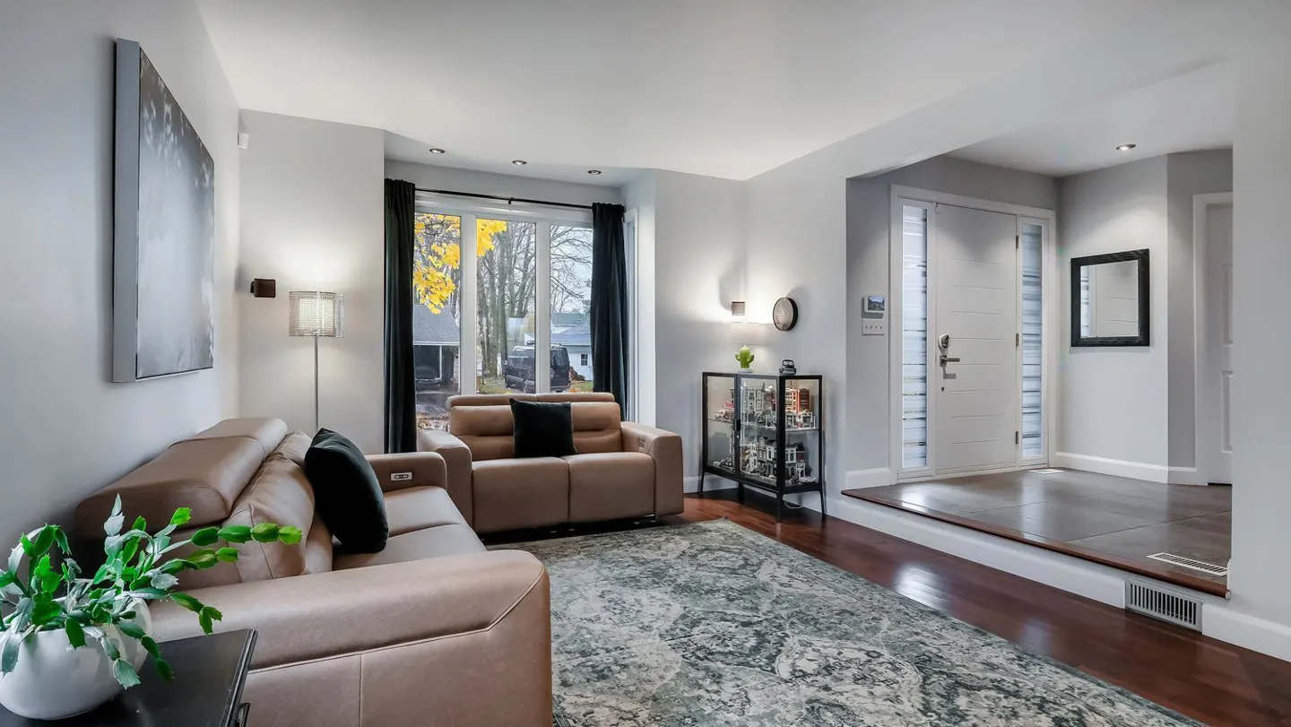 Living room with tan leather sofas, black pillows, and a patterned rug. A window with black curtains overlooks a yard. A white front door is visible.