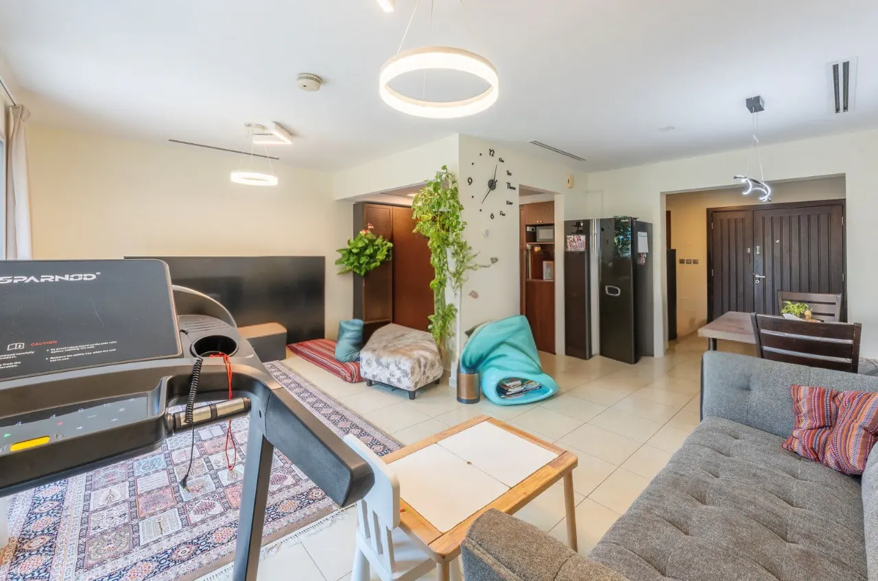 A bright living room with a treadmill, gray sofa, and dining area. A rug and plants add color to the neutral space.