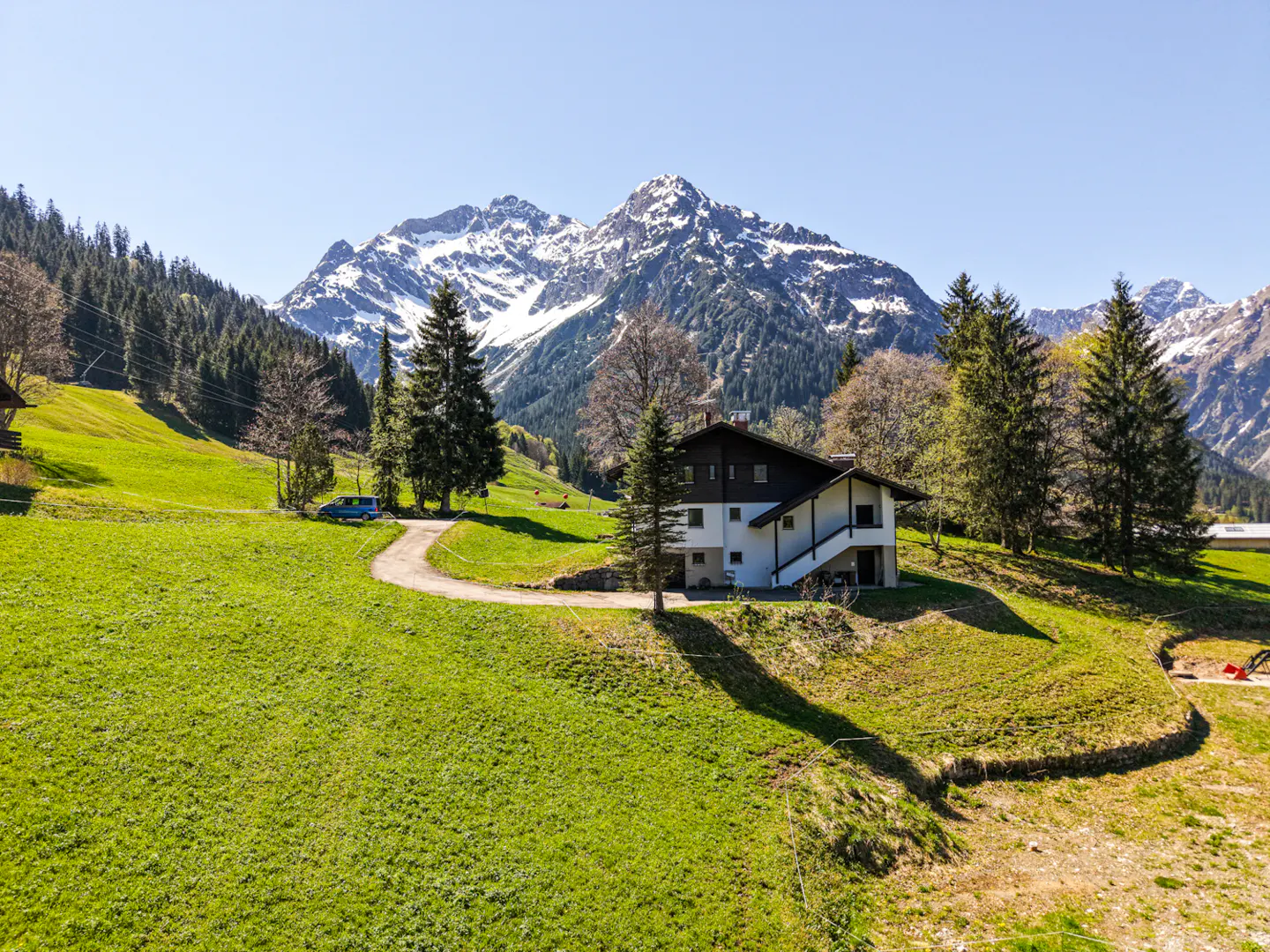 A white house with a dark roof sits on a green hill, with snow-capped mountains in the background.