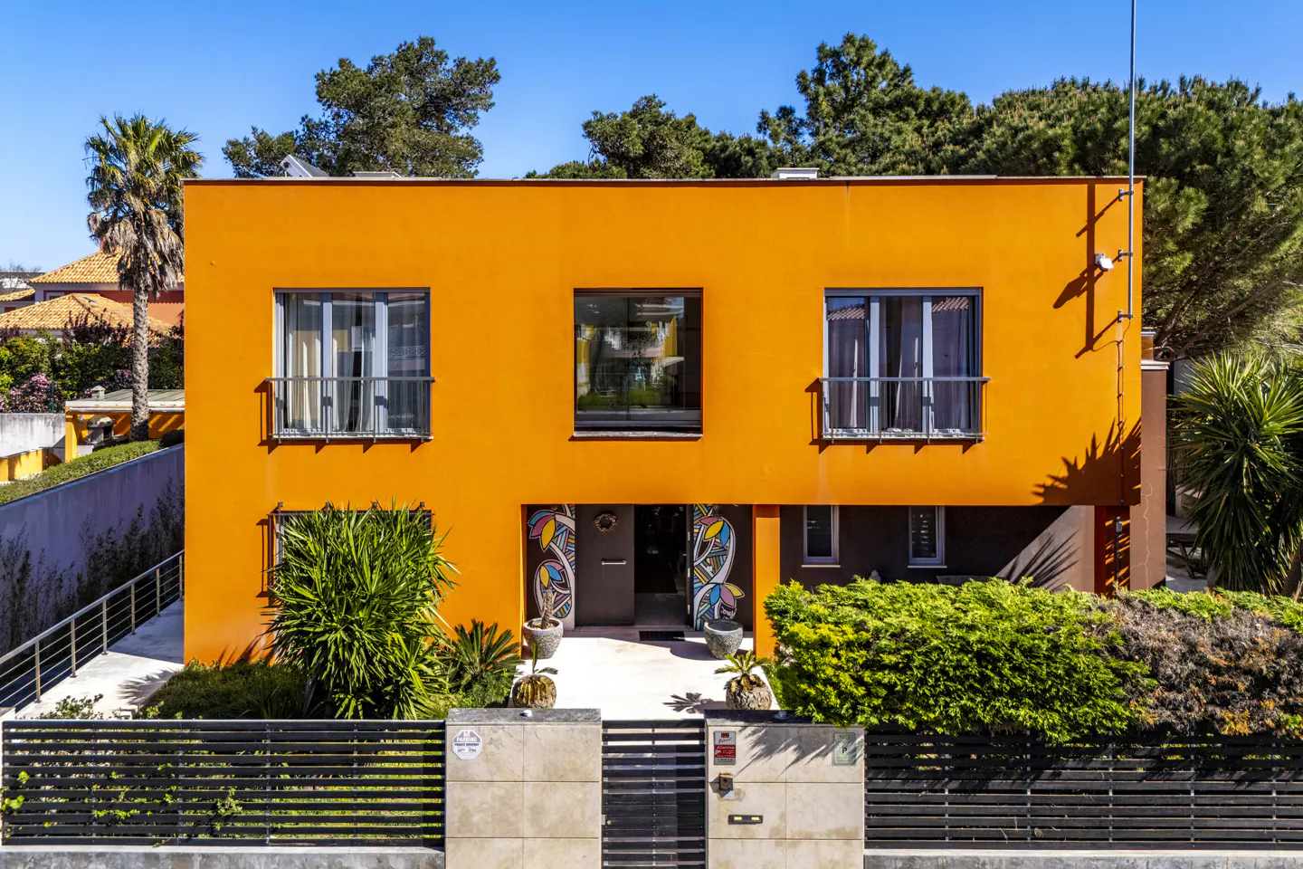 Two-story orange house with a flat roof, black fence, and green bushes in front. The front door has colorful art on either side.
