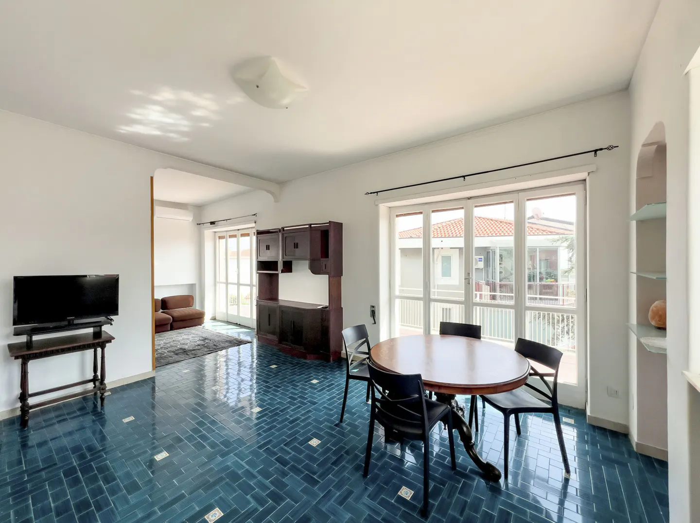 Bright living room with blue tile floor, round wood table with black chairs, and a dark wood cabinet. A TV is on a stand in the background.