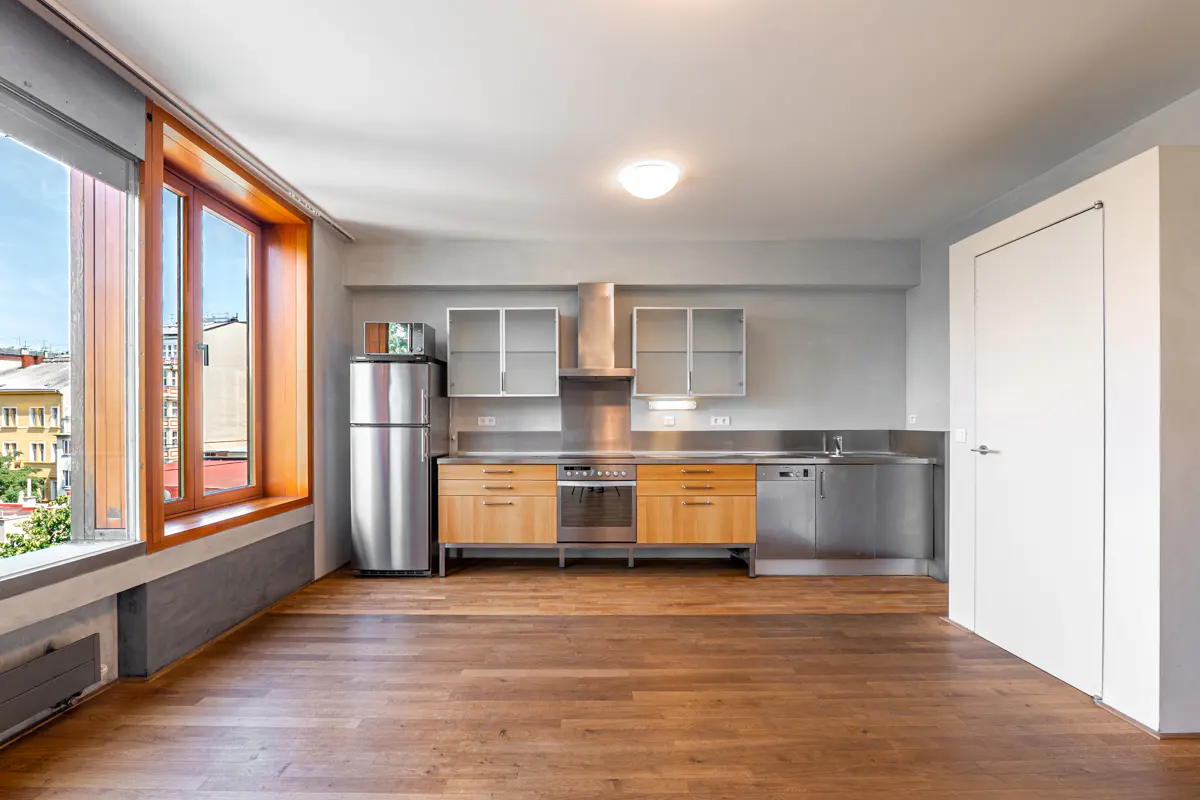 Bright, empty kitchen with wood floors, stainless steel appliances, and a large window with a view of buildings.