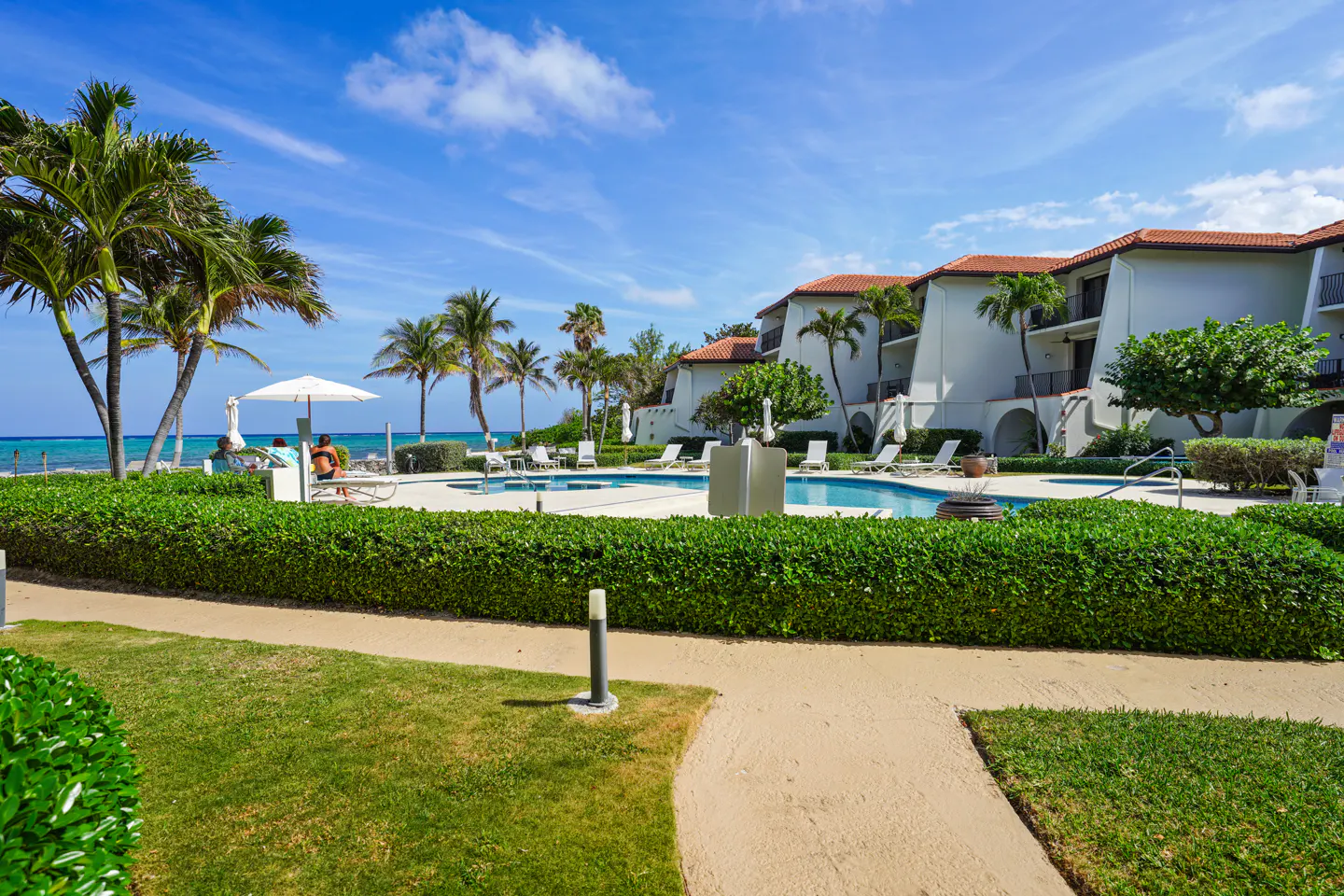 Resort view with a pool, white buildings with red roofs, palm trees, and a blue sky.