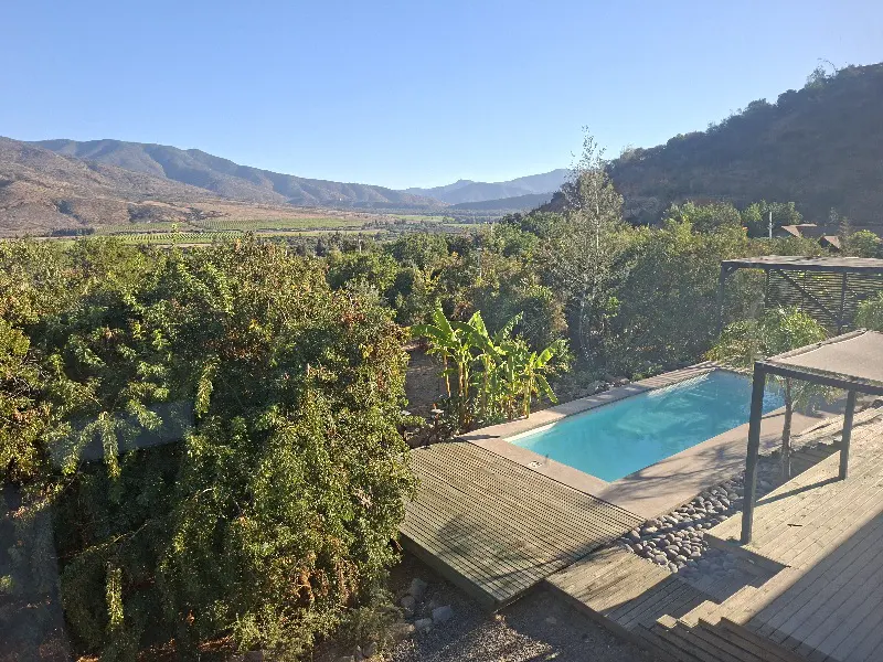 A landscape view of a pool with blue water, a wooden deck, and lush green trees with mountains in the background.