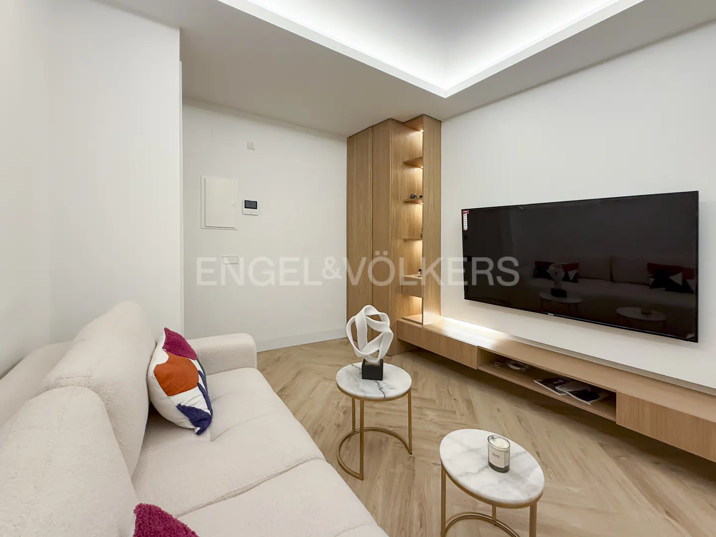 Living room with a white sofa, colorful pillows, two marble tables, a TV, and a wooden cabinet on a herringbone wood floor.