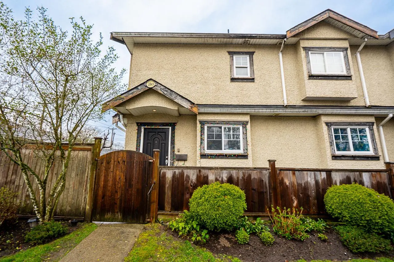 Tan townhouse with a dark door, white windows, and a brown wooden fence with green bushes in front.