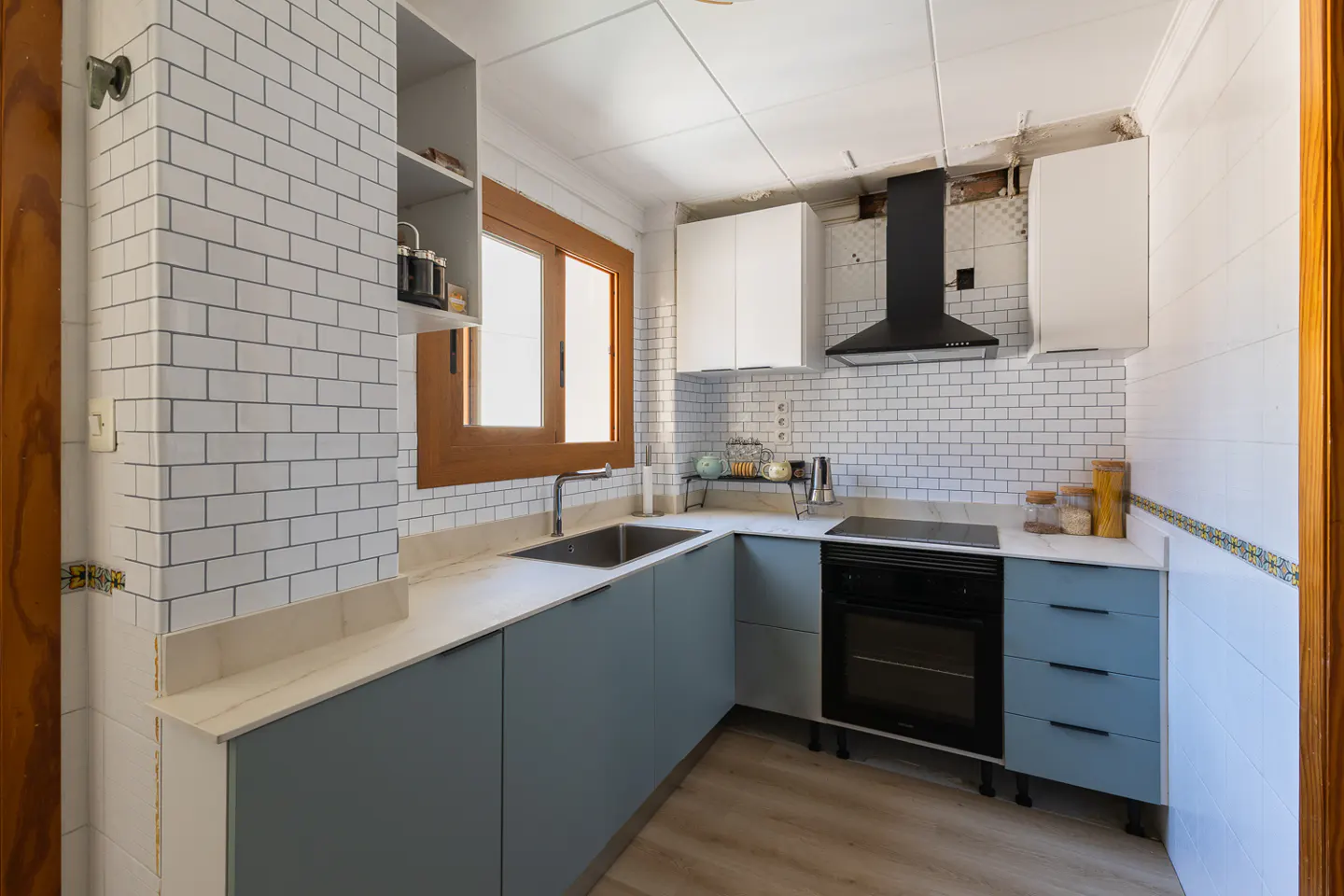 Bright kitchen with blue cabinets, white subway tile, and a black range hood. A wooden window frame adds warmth to the space.