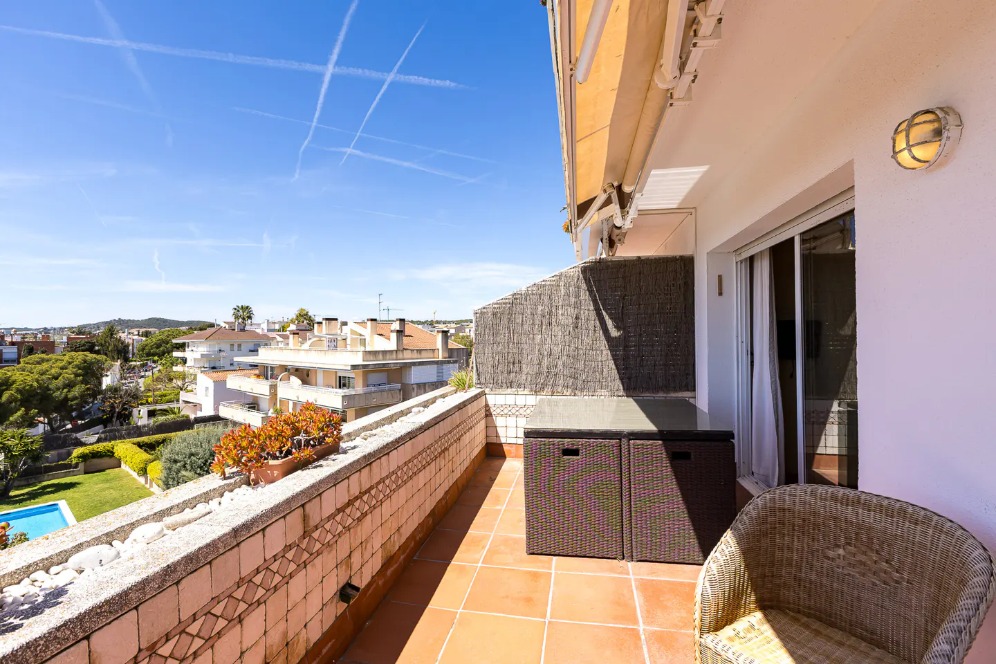 Balcony view with wicker chair, brown storage boxes, and tiled floor. A view of houses, green trees, and a blue sky is in the background.