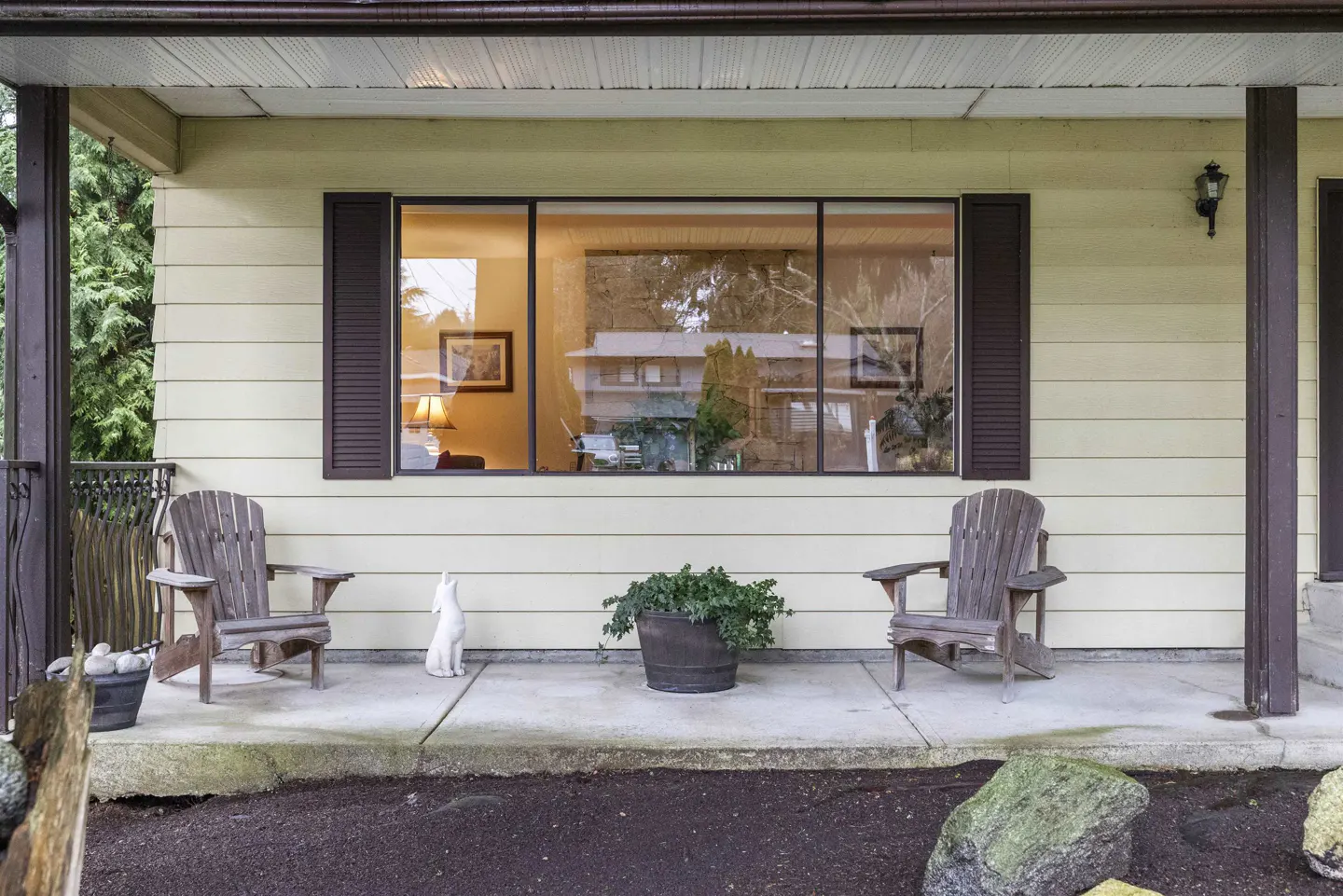 Front porch of a yellow house with two wooden chairs, a potted plant, and a white rabbit statue. A large window reflects the house across the street.