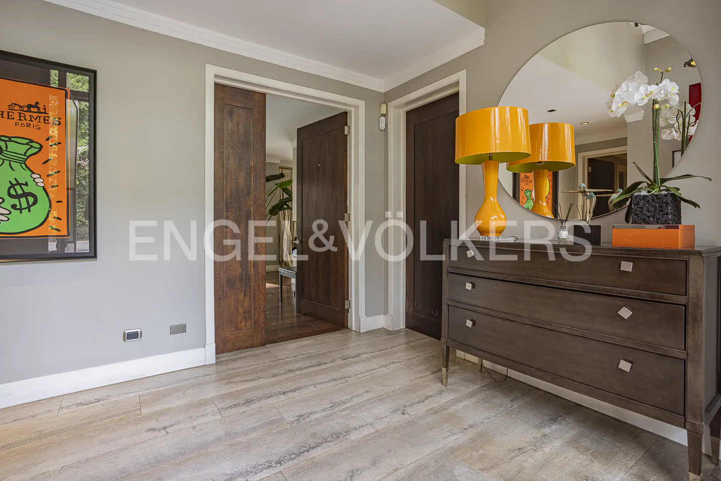 Entryway with light wood floors, dark wood dresser, bright orange lamp, round mirror, and Hermes art on a gray wall.