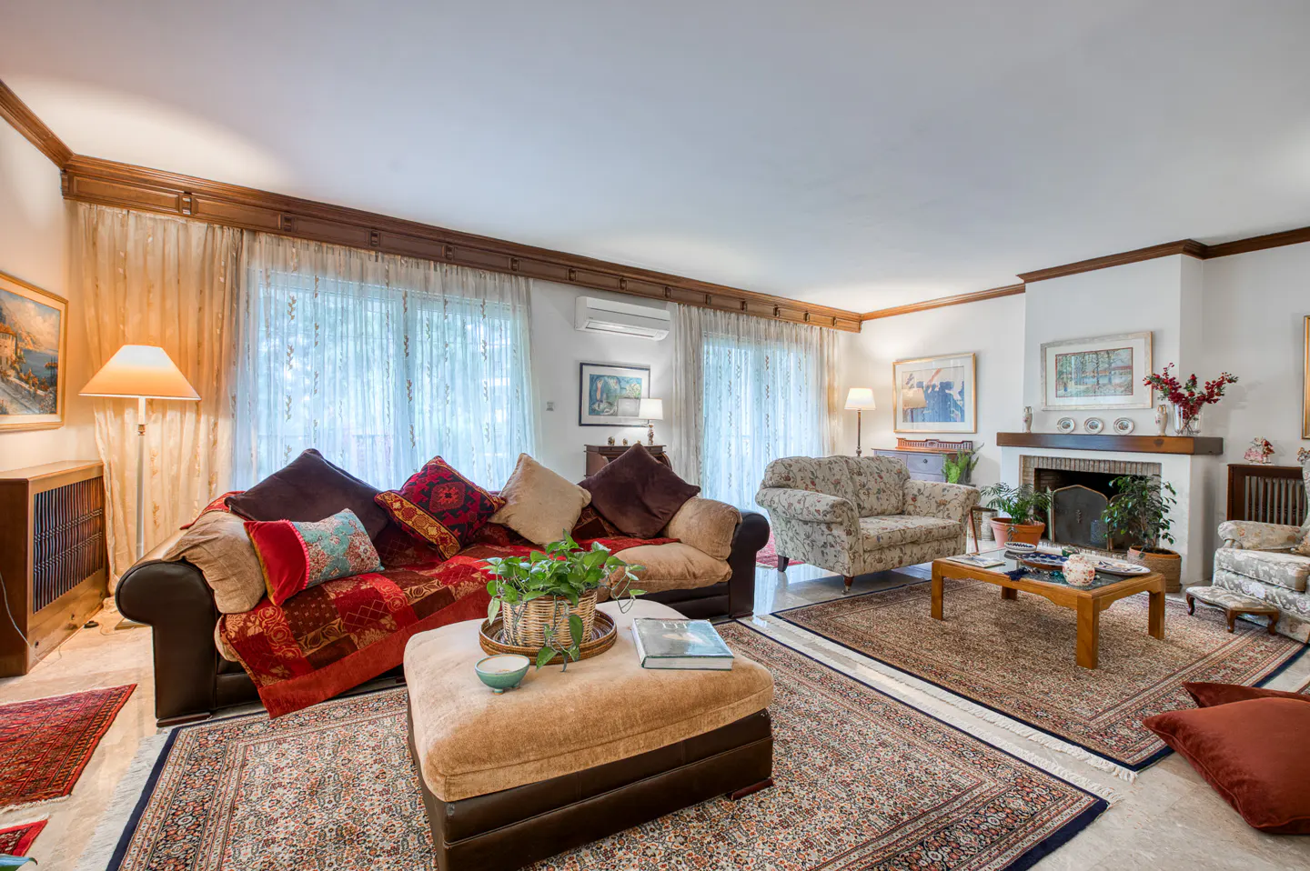 A living room with a brown sofa, patterned pillows, and a tan ottoman on a patterned rug. A fireplace and floral armchair are in the background.