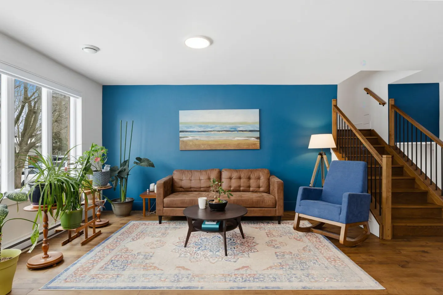 Living room with blue accent wall, brown leather sofa, blue rocking chair, and wooden staircase. A large window lets in natural light.