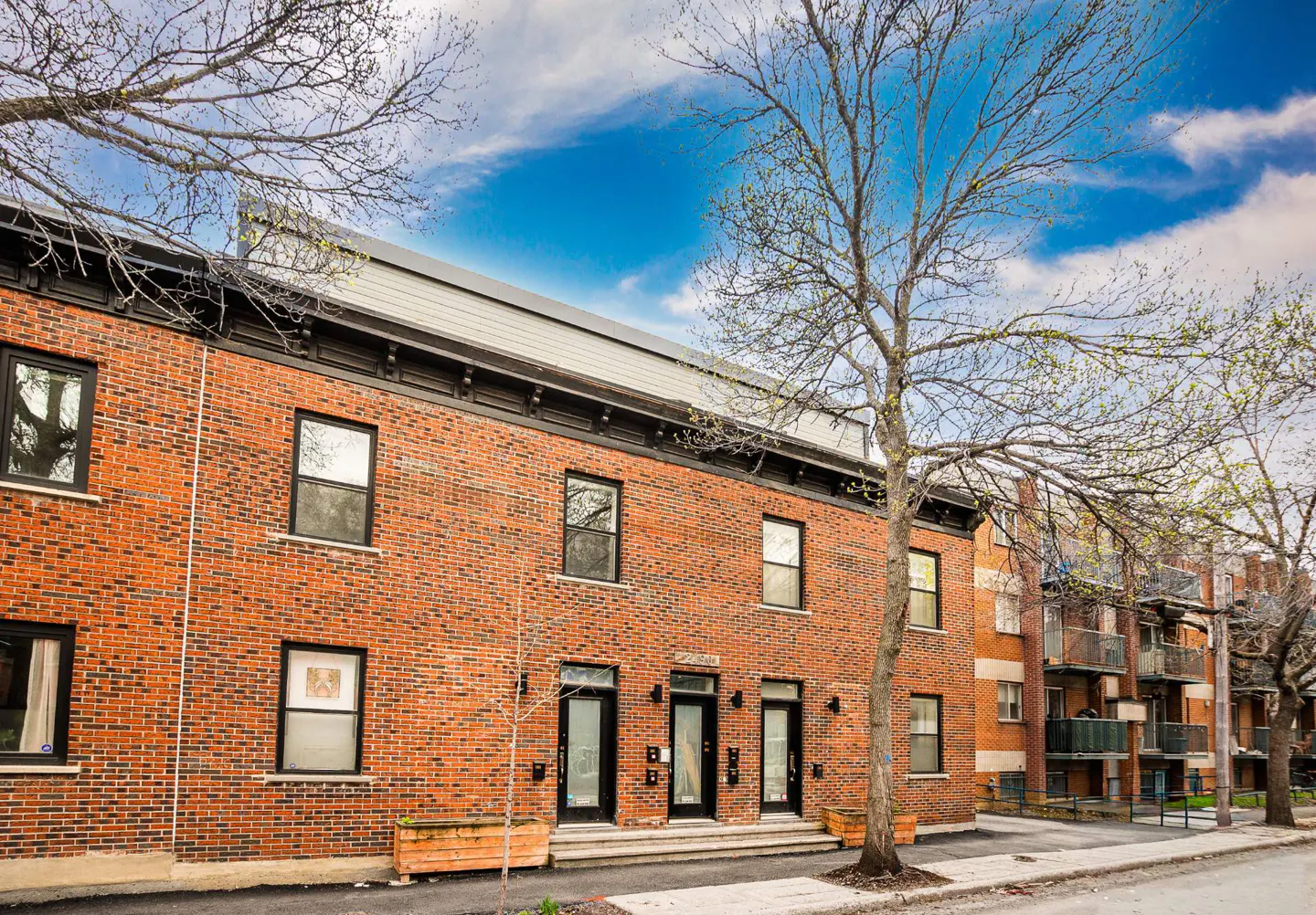 Red brick townhouses with black trim and doors line a street under a blue sky with bare trees.