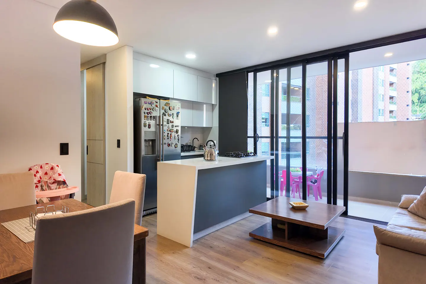 A modern apartment interior with a kitchen island, dining table, and balcony. The color scheme is neutral with pops of color from the balcony furniture.