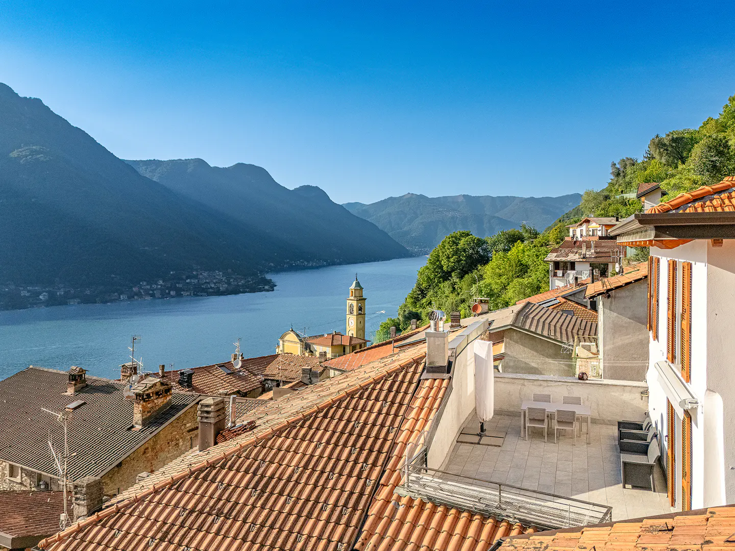 Scenic view of Lake Como, Italy, with terracotta rooftops, a church steeple, and mountains under a clear blue sky.