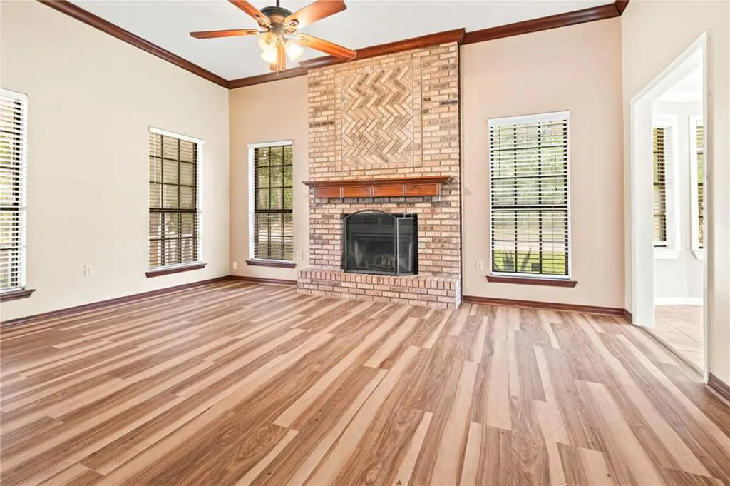 Living room with wood floors, brick fireplace, and ceiling fan. Windows with blinds flank the fireplace. Cream colored walls and dark wood trim.