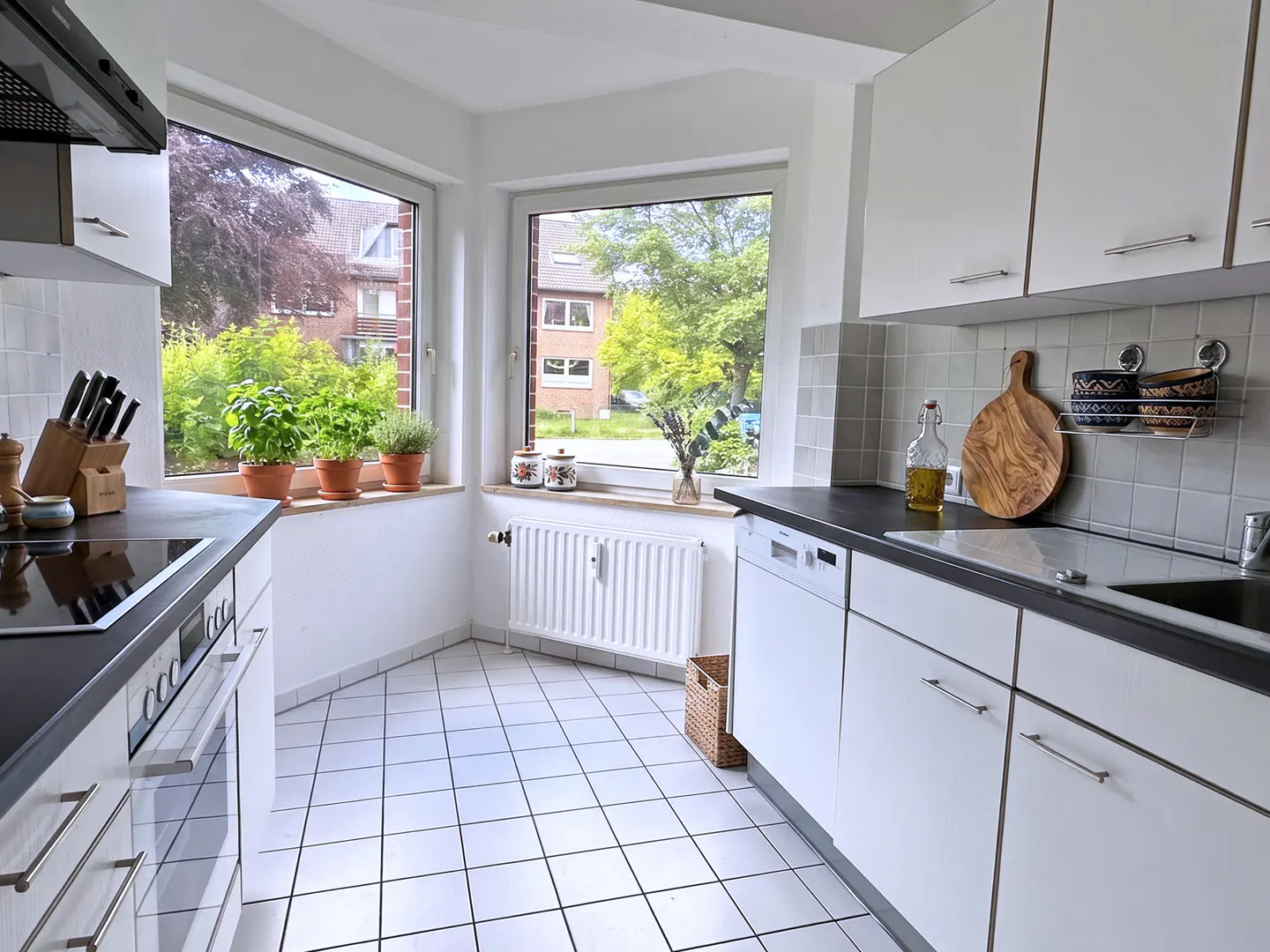 Bright kitchen with white cabinets, black countertops, and white tiled floor. Windows overlook greenery. Herbs in pots sit on the windowsill.