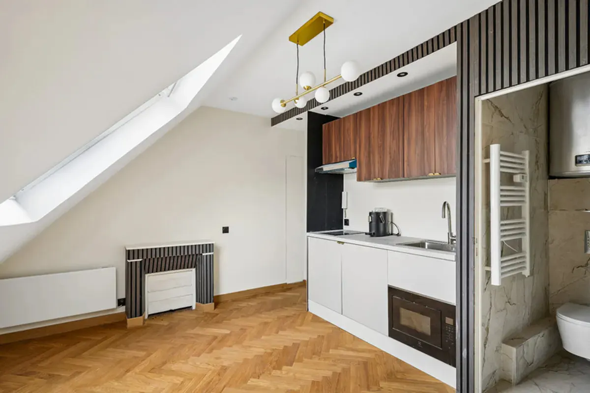 Modern attic kitchen with wood cabinets, white counters, herringbone floors, and an adjacent marble-tiled bathroom.