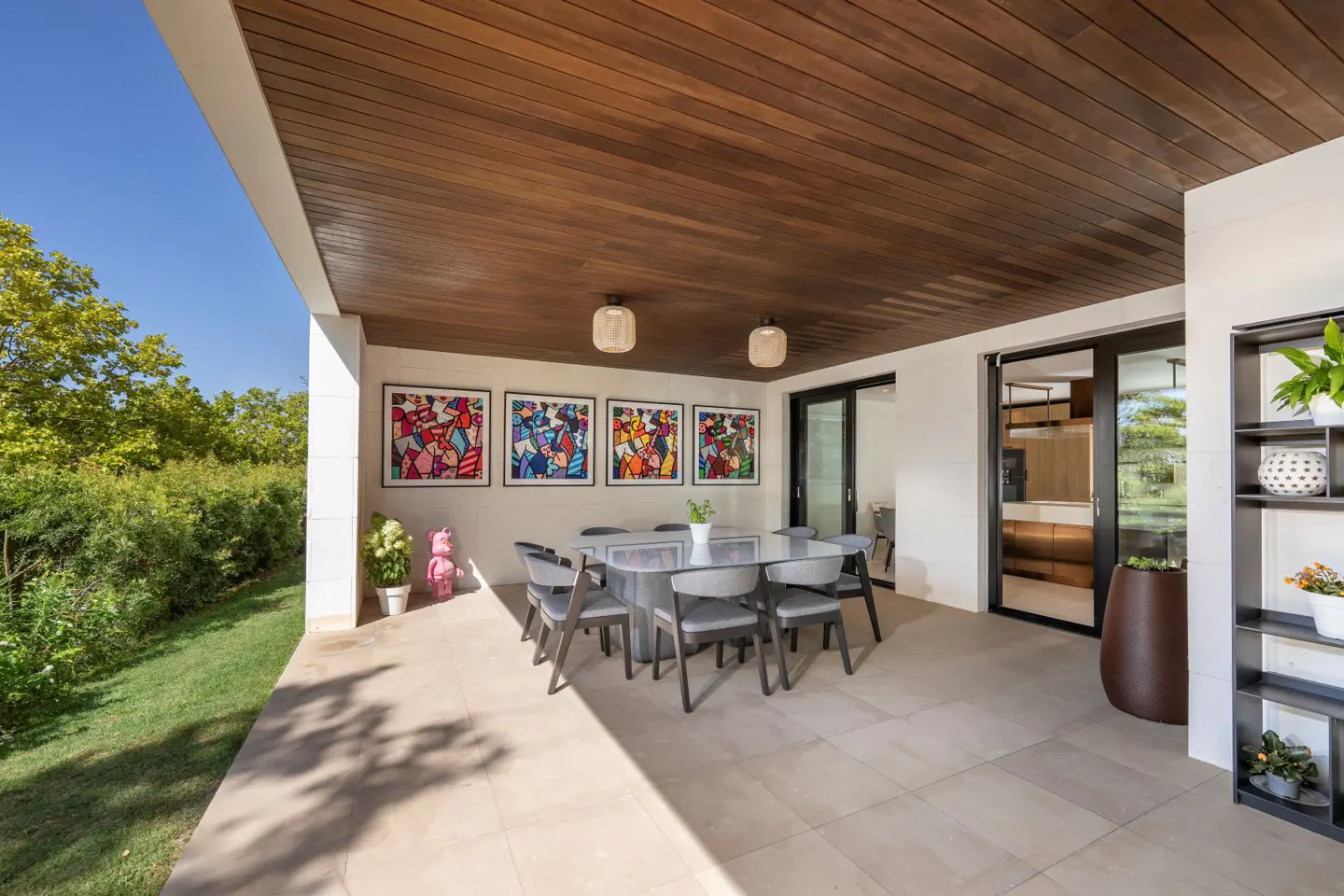 Covered patio with a dining table, chairs, and colorful artwork. A pink bear figurine and plants add charm. Sliding doors lead inside.