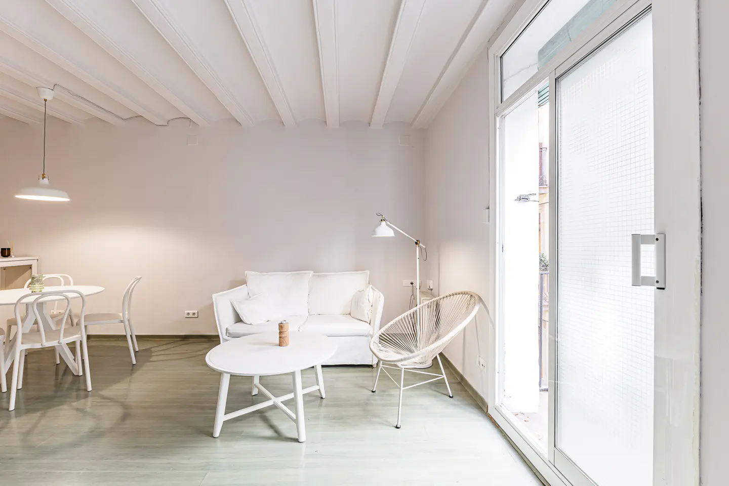 Bright, minimalist living room with white furniture, including a sofa, table, chairs, and a woven chair near a sliding glass door.