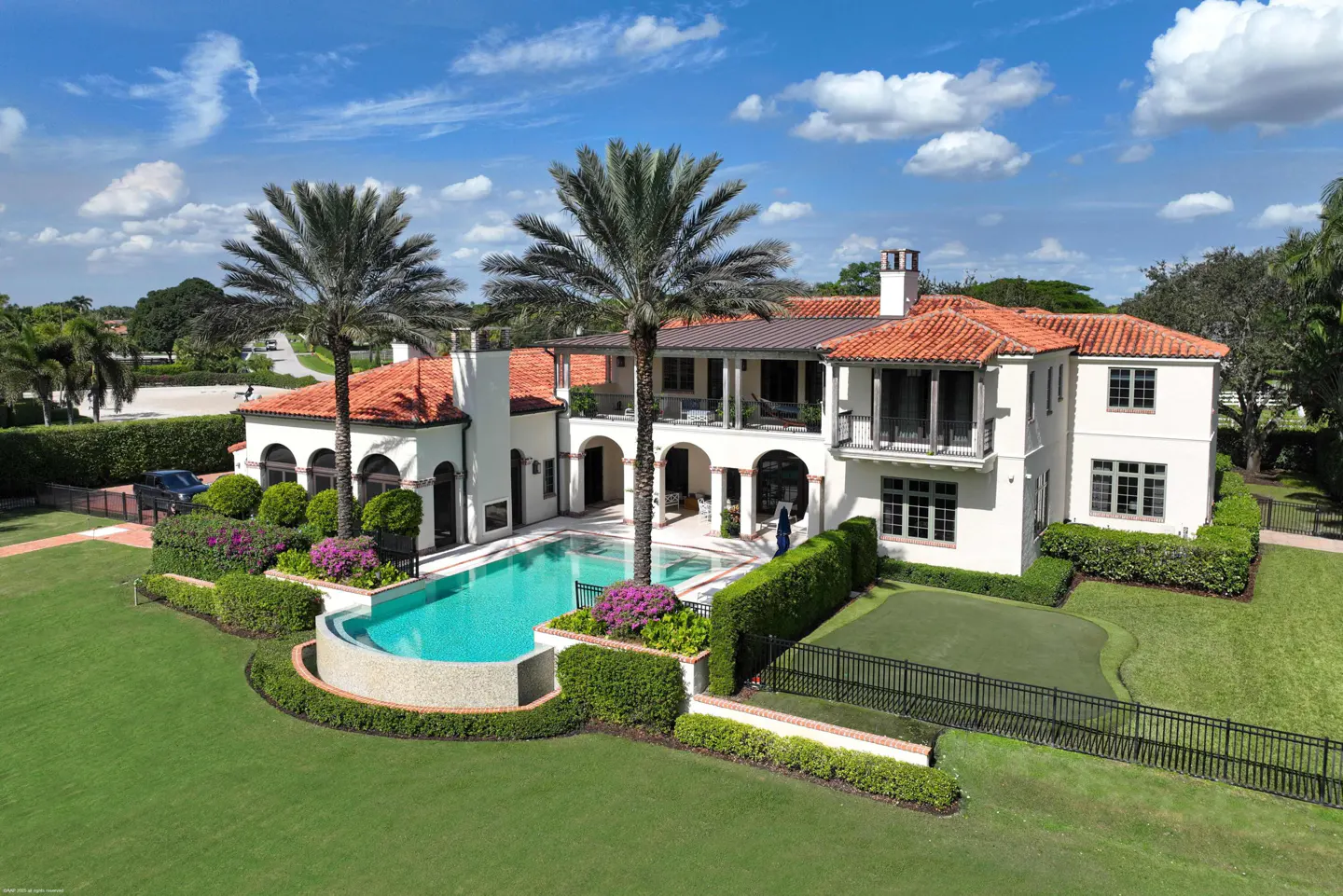 Aerial view of a large, white, two-story house with a red tile roof, a pool, palm trees, and a green lawn.