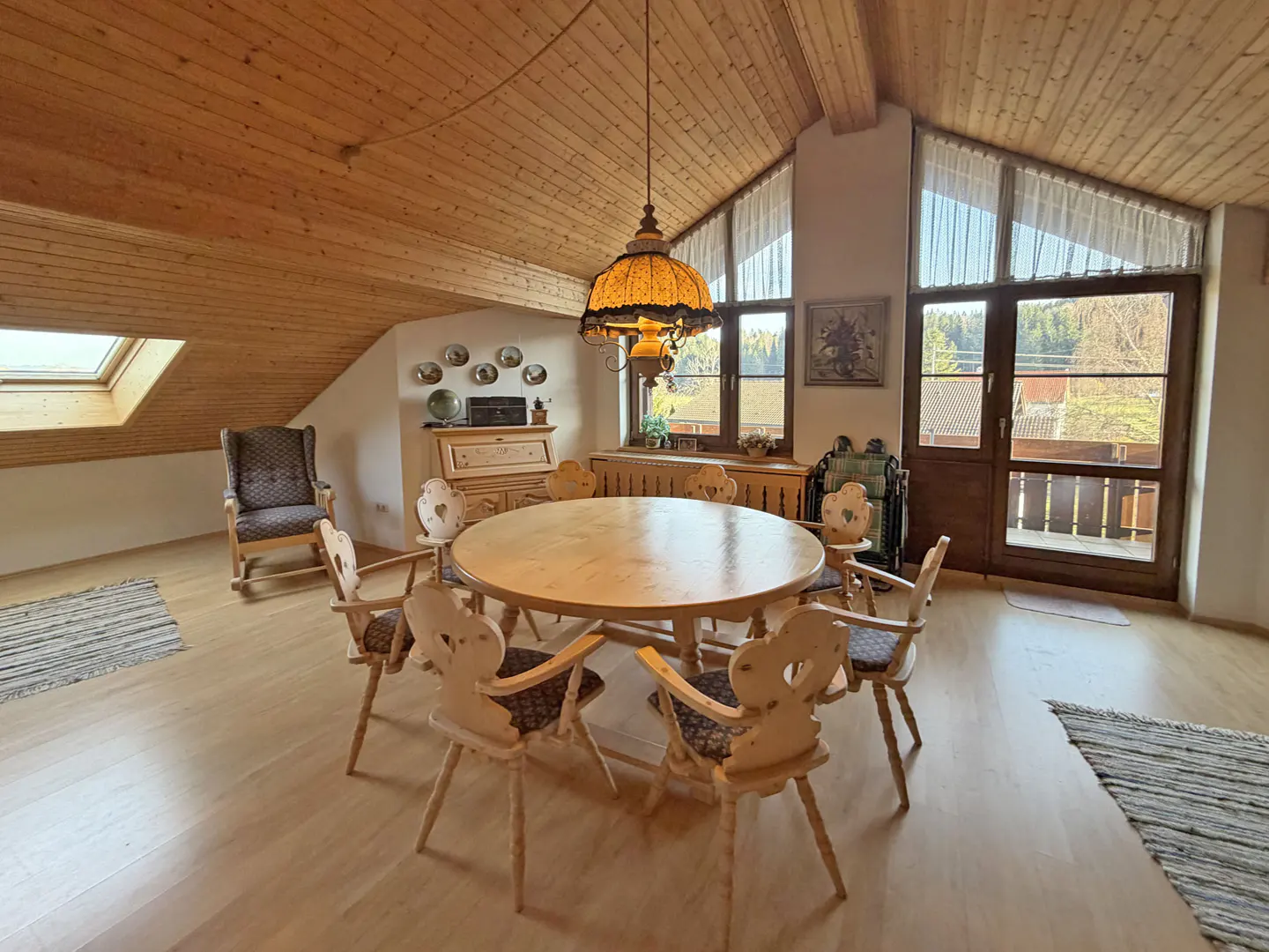 Attic dining room with wood-paneled ceiling, round table with heart-backed chairs, and windows overlooking a wooded landscape.