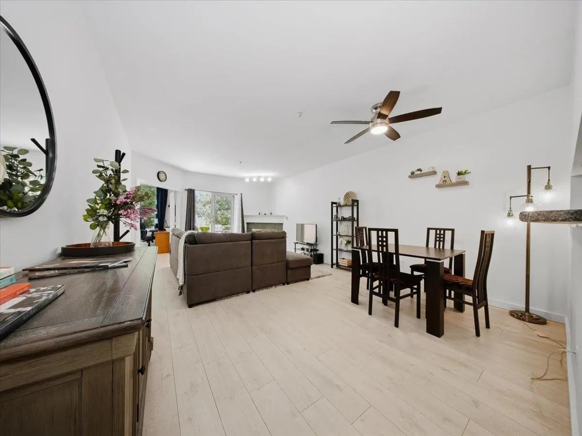Bright, open-concept living space with light wood floors, a brown sectional sofa, and a dark wood dining set. A ceiling fan and large windows add to the airy feel.