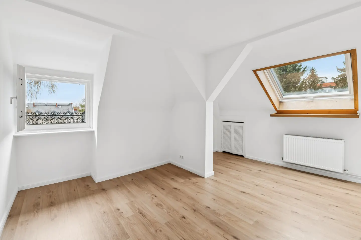 Bright, empty attic room with light wood floors, white walls, and two windows. One window is a skylight. A white radiator is on the wall.