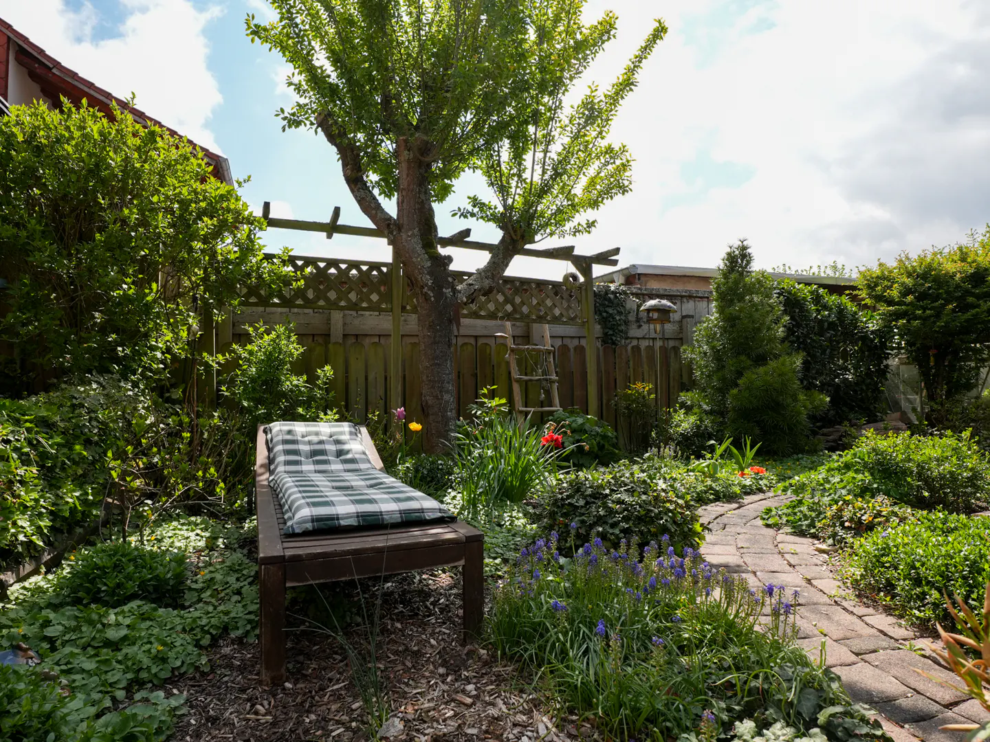 A wooden lounge chair with a plaid cushion sits in a lush garden with a stone path, mature tree, and wooden fence.