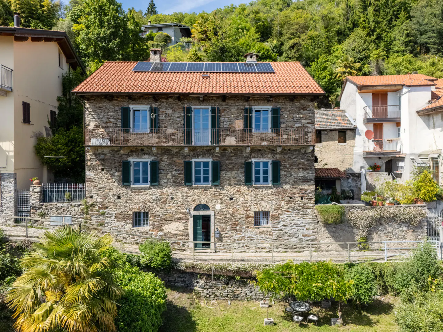 Two-story stone house with a red tile roof and solar panels, green shutters, and a small garden.