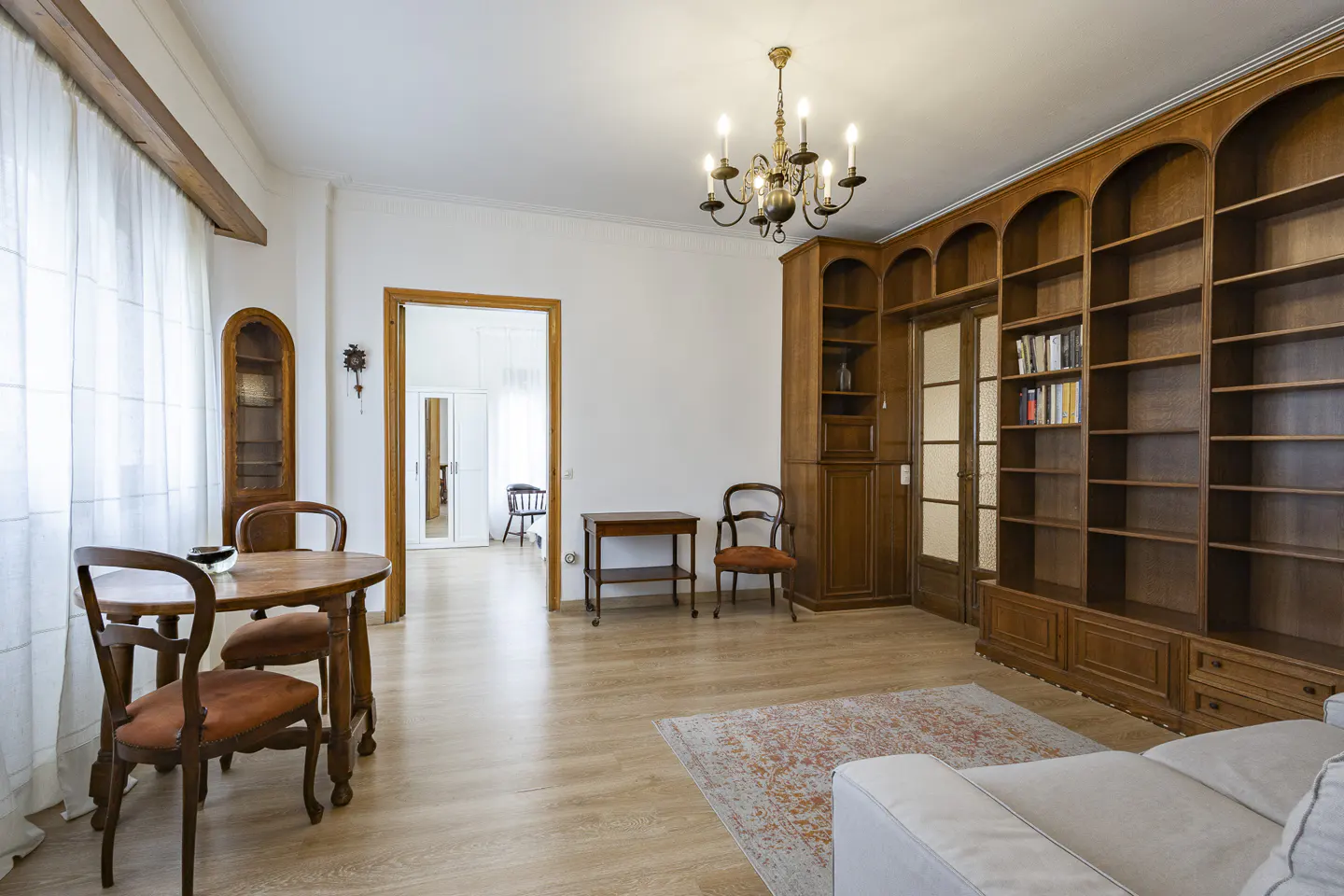A living room with wood floors, a large wooden bookcase, and a round wooden table with chairs. A chandelier hangs from the ceiling.